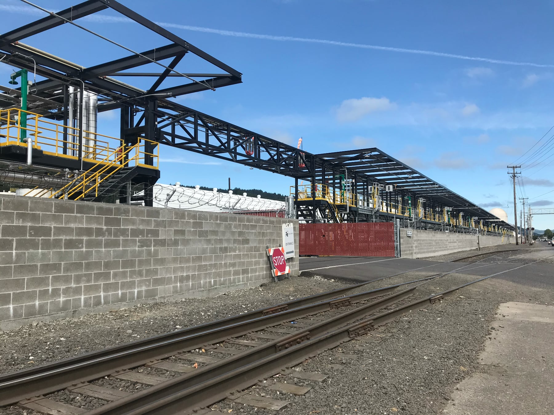 The front gate of Zenith Energy. A cement block wall recedes into the distance behind which are the platforms and piping that are part of the facilities unloading apparatus.